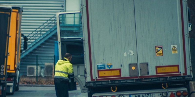 Motorista usando colete de alta visibilidade descarrega uma carga ao lado de um caminhão em uma área industrial.