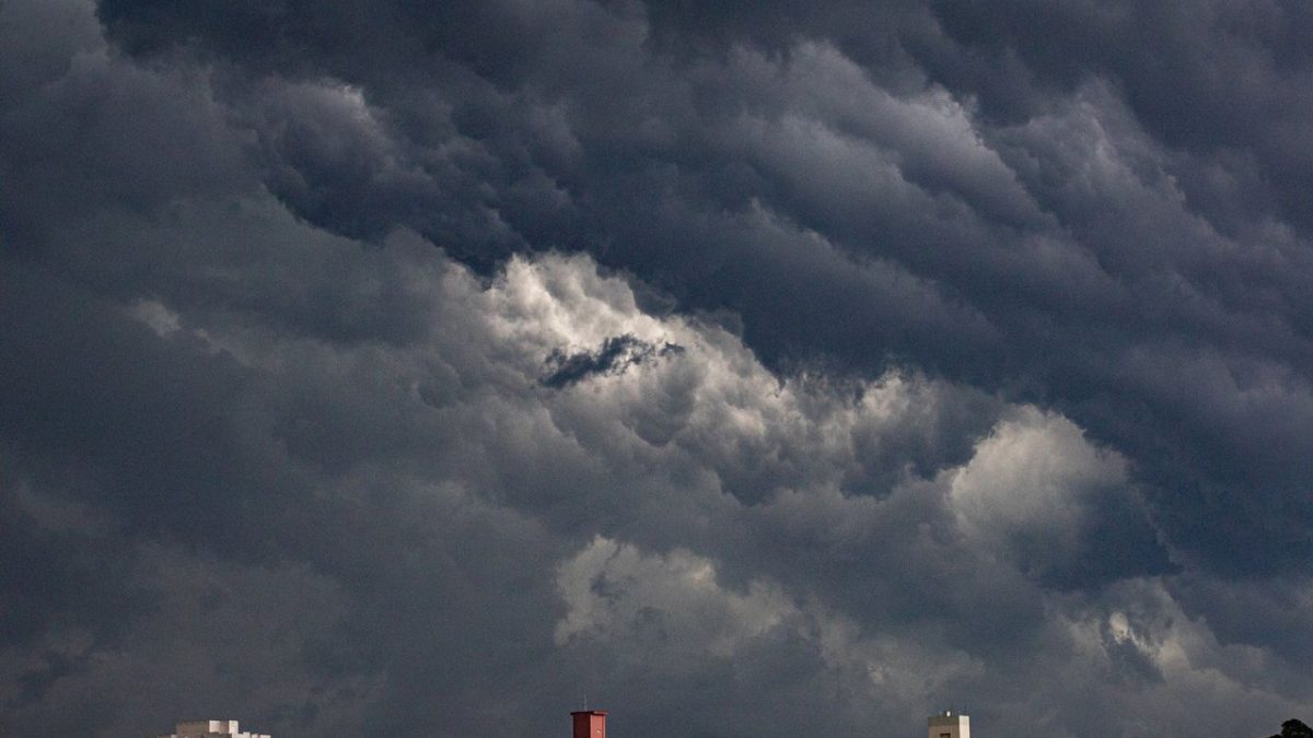 Nuvens escuras se acumulam sobre prédios de uma área urbana, indicando a aproximação de uma tempestade.