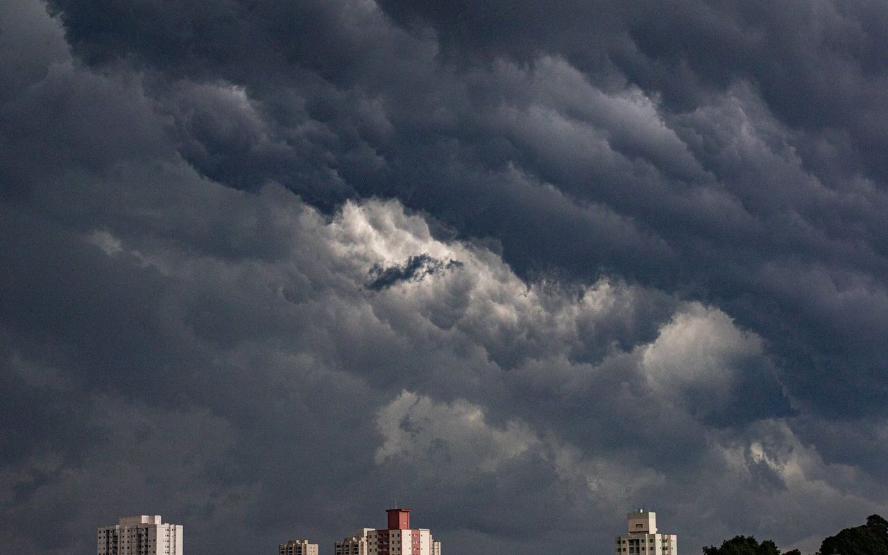 Nuvens escuras se acumulam sobre prédios de uma área urbana, indicando a aproximação de uma tempestade.